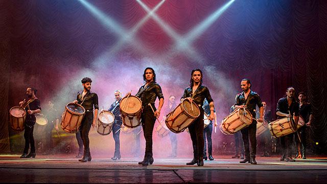 a group of men stand in formation on a stage lit with spotlights and fog while holding large drums